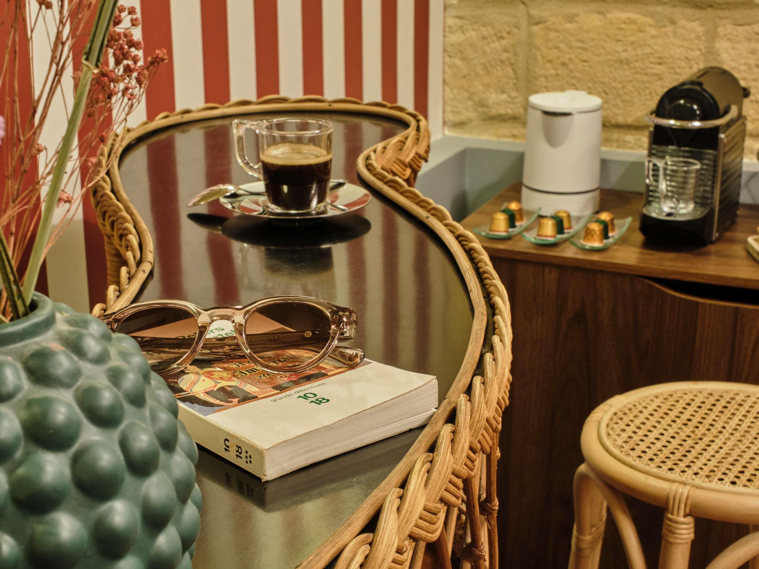 A wicker-framed table with a book, a pair of sunglasses, a coffee cup, and a coffee maker.