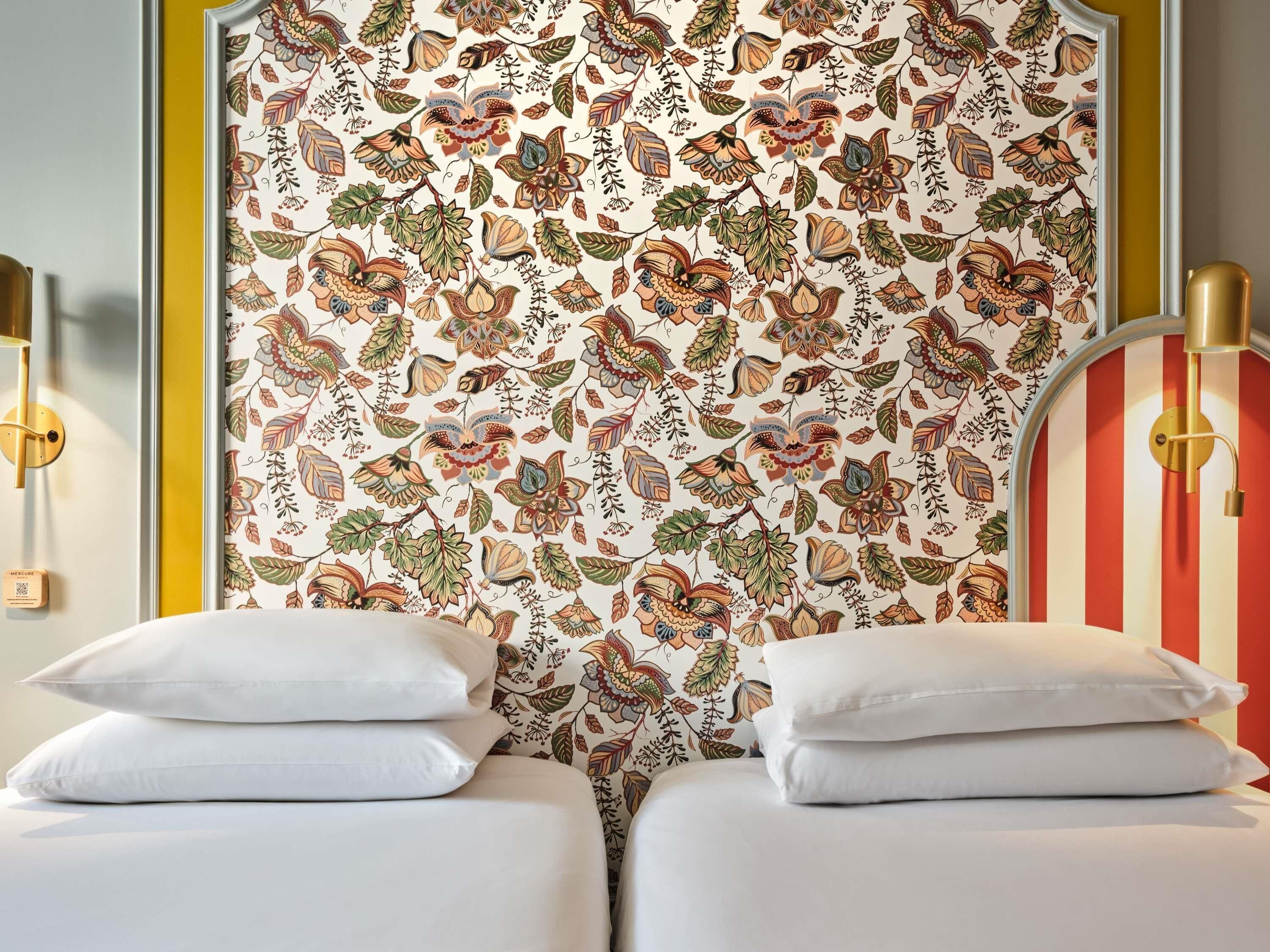 A hotel room with a floral-patterned headboard, two white beds with pillows, and a striped accent wall.