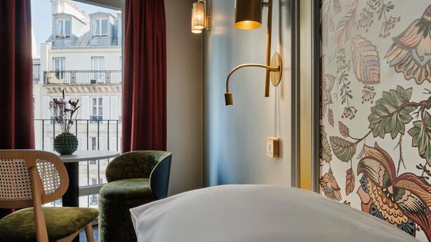 Une chambre d’hôtel avec un lit, une chaise, une lampe fixée au mur et une fenêtre avec des rideaux.