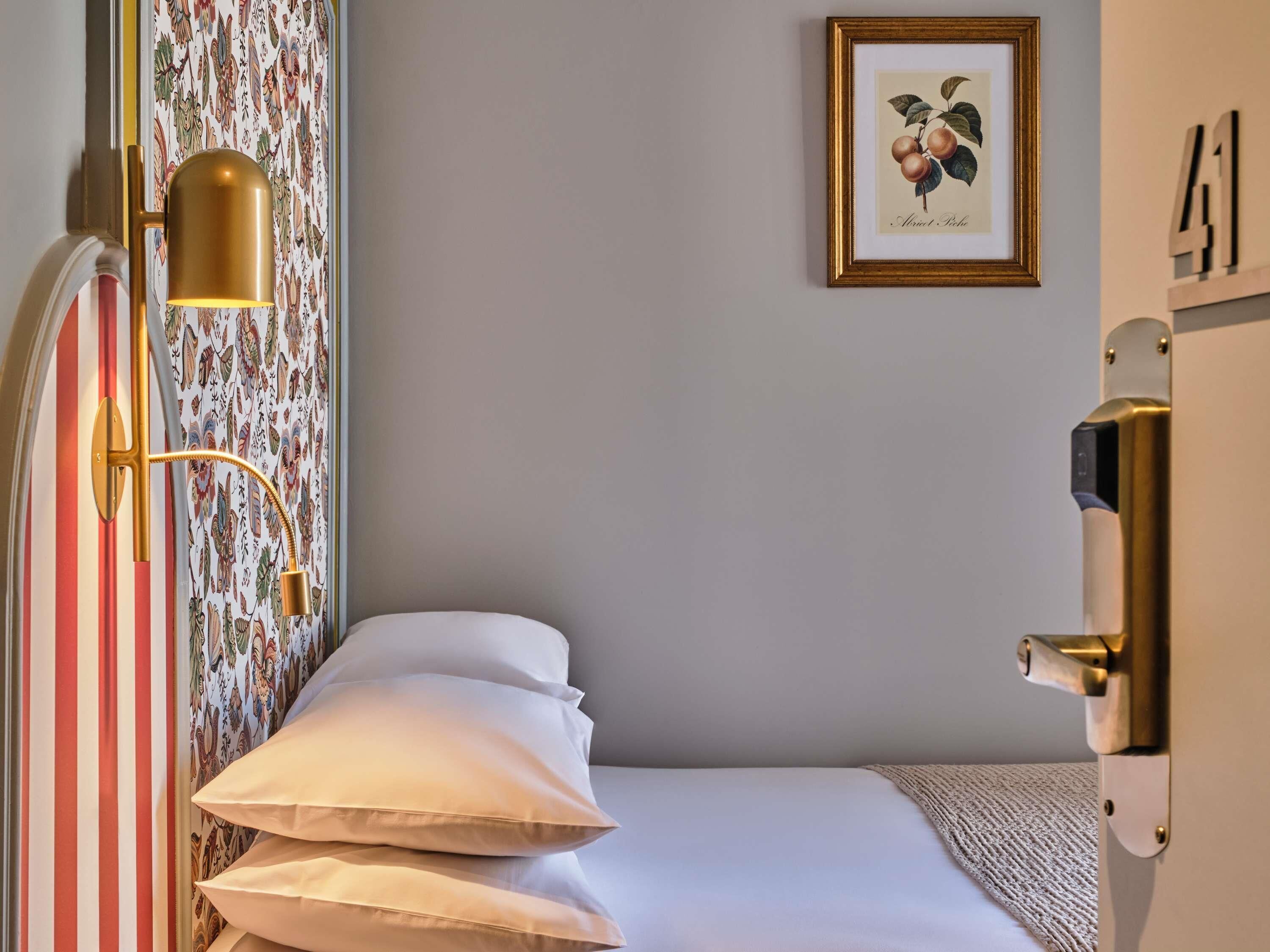 Ett hotellrum med en säng, en vägglampa, en inramad botanisk illustration och en dörr med ett handtag.