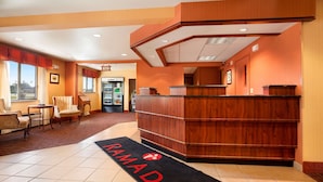 A reception area with a wooden reception desk, a black carpet with 'RAMADA' logo, and a seating area with chairs and a small table. - Ramada by Wyndham Wisconsin Dells (Baraboo)