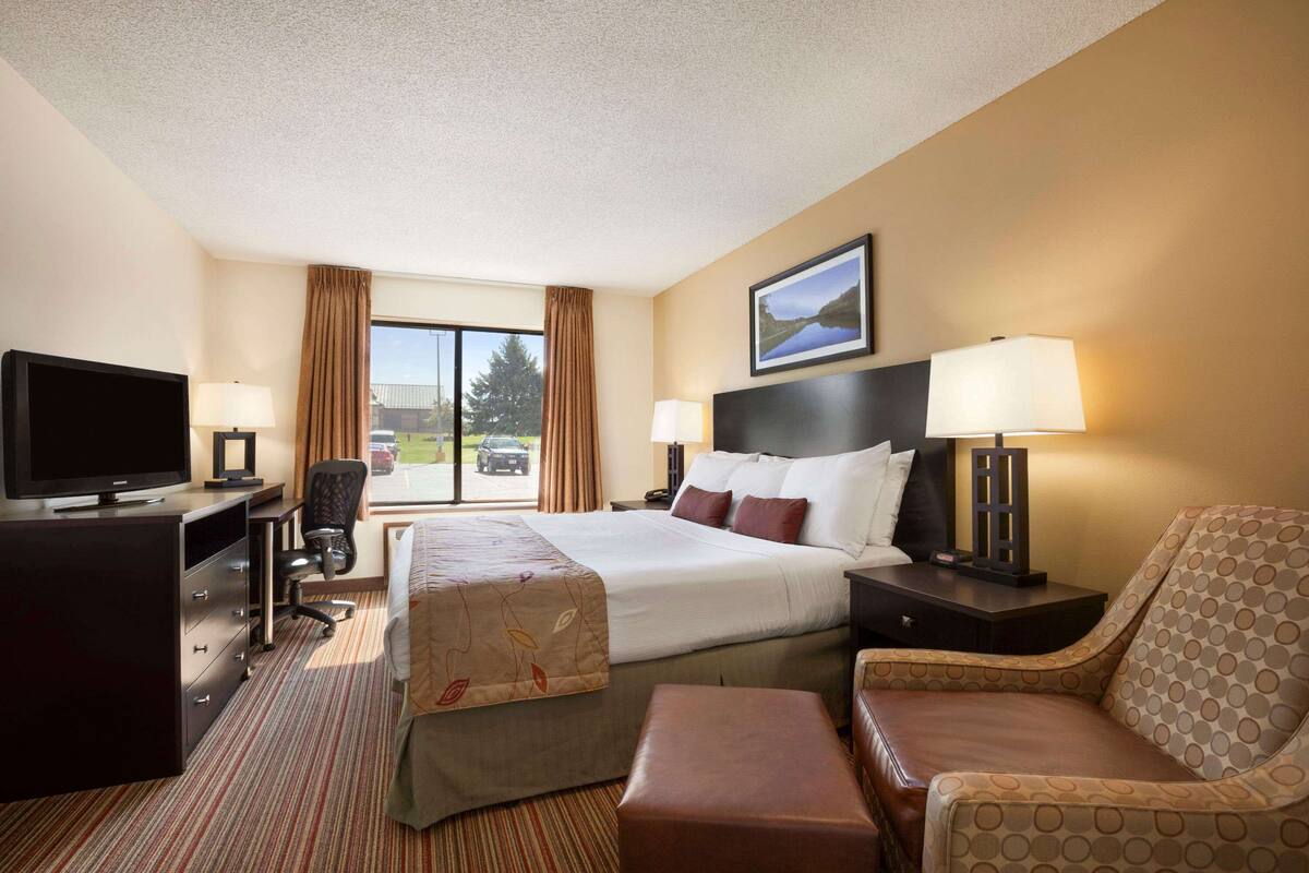 A hotel room with a large bed, two bedside tables with lamps, a desk with a TV, a chair, and a view of the outdoors.