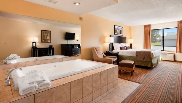 A hotel room with a large bathtub, a bed with pillows, a chair, a desk, a television, and a window with curtains. - Ramada by Wyndham Wisconsin Dells (Baraboo)