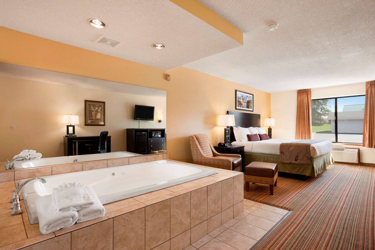 A hotel room with a large bathtub, a bed with pillows, a chair, a desk, a television, and a window with curtains.