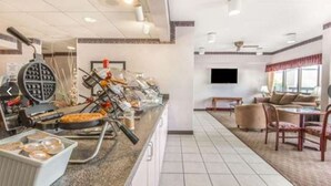 A hotel breakfast area with a buffet setup, a seating area with a TV, and a window providing natural light. - Baymont by Wyndham Topeka (Topeka)