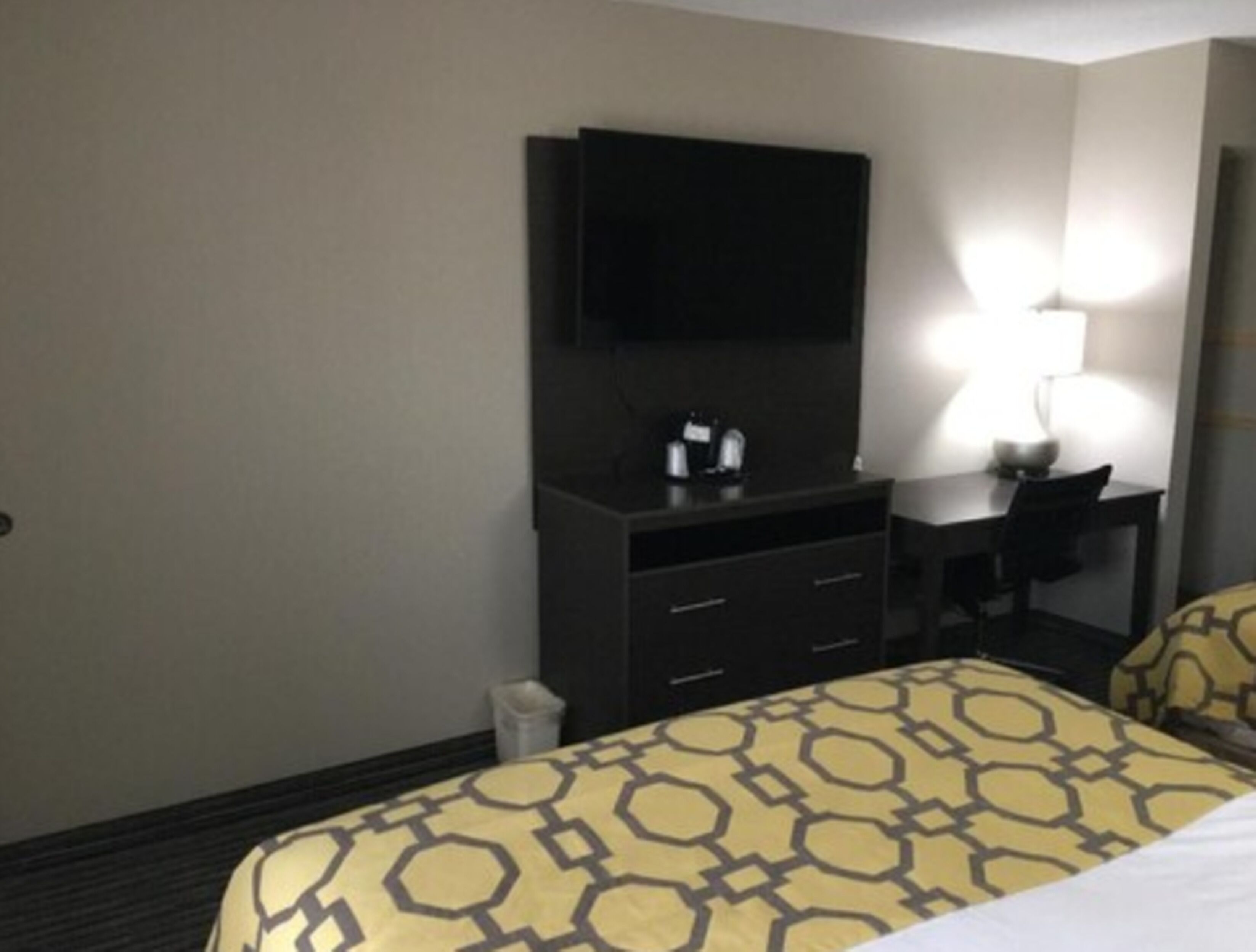 A hotel room with a bed, a desk, a chair, a TV mounted on the wall, and a lamp on the desk.