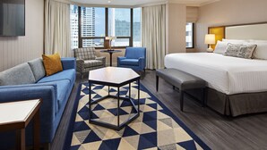 A hotel room with a bed, a sofa, a chair, a small table, and a view of the cityscape through large windows.