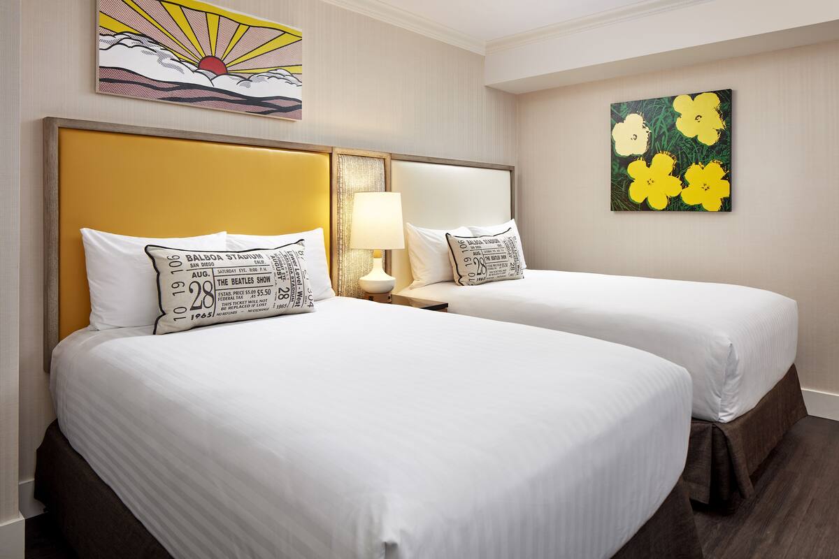 A hotel room with two beds, each with white bedding and decorative pillows. There is a nightstand with a lamp between the beds.