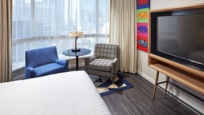 A hotel room with a large bed, a blue armchair, a plaid armchair, a small table with a lamp, a TV on a wooden stand, and a city view through large windows.