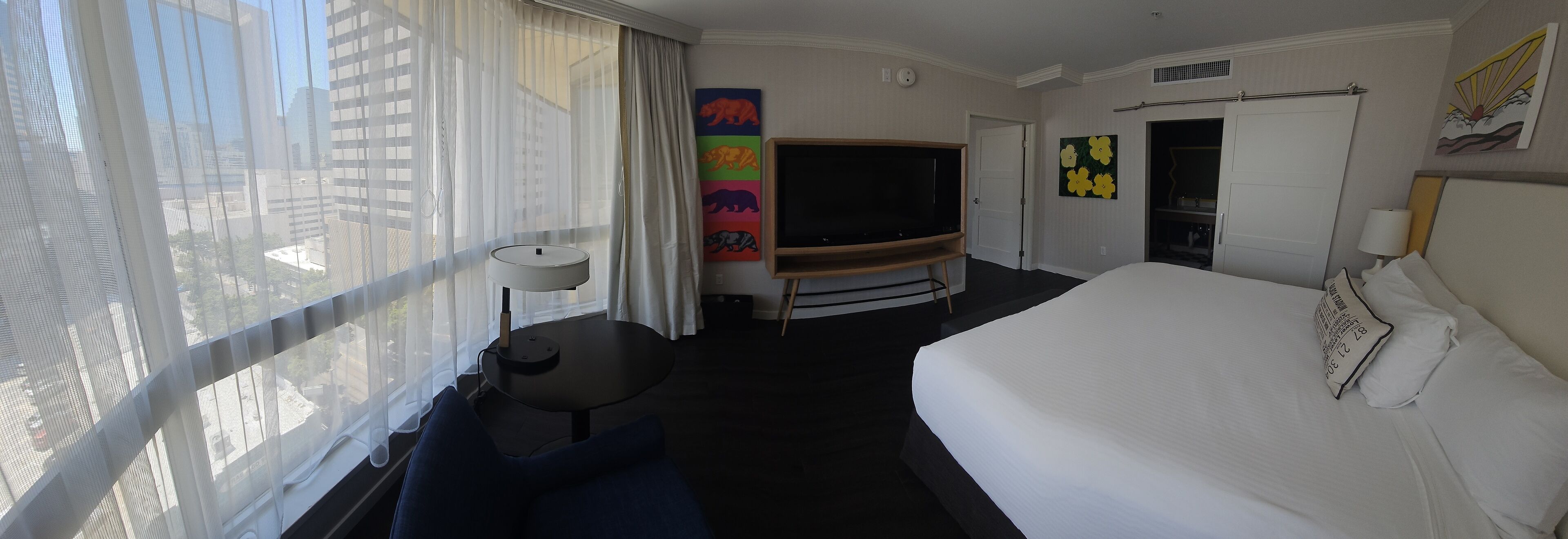 Habitación de hotel con una cama grande, televisión, escritorio y ventana con cortinas translúcidas.