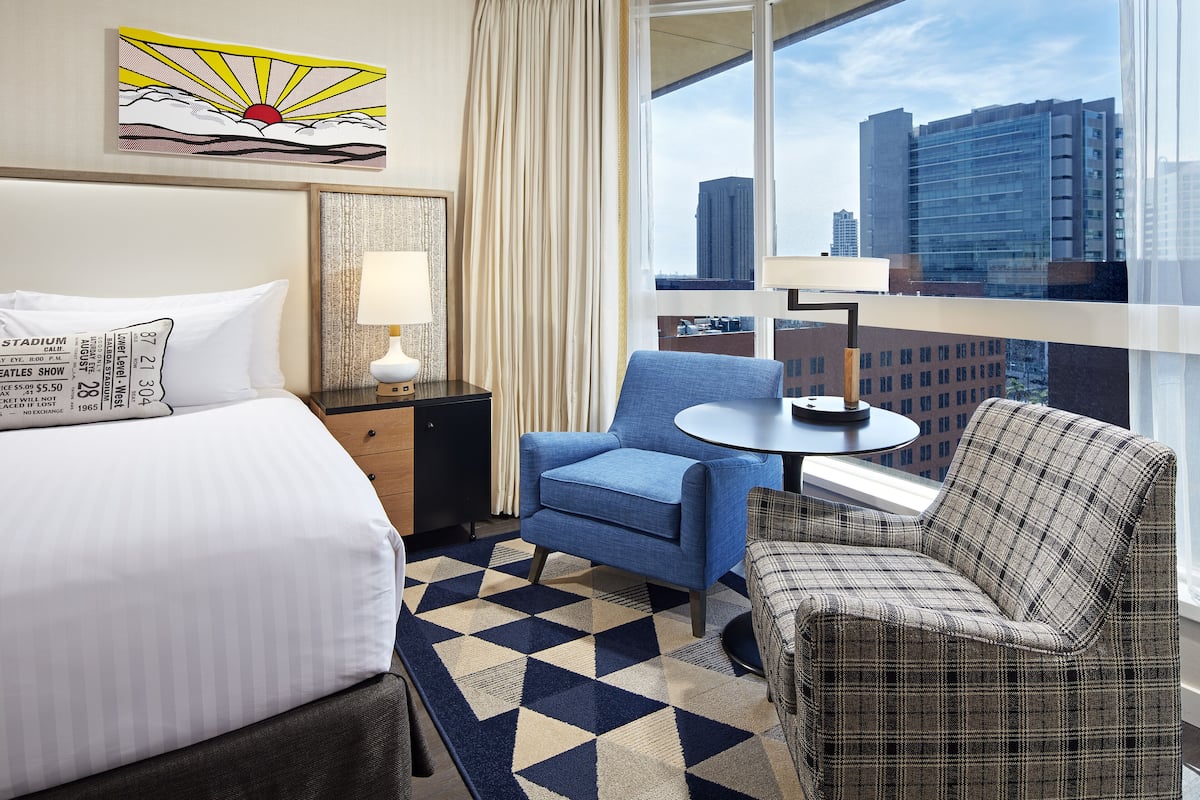 A hotel room with a bed, a blue armchair, a plaid armchair, a small table, and a city view through large windows.