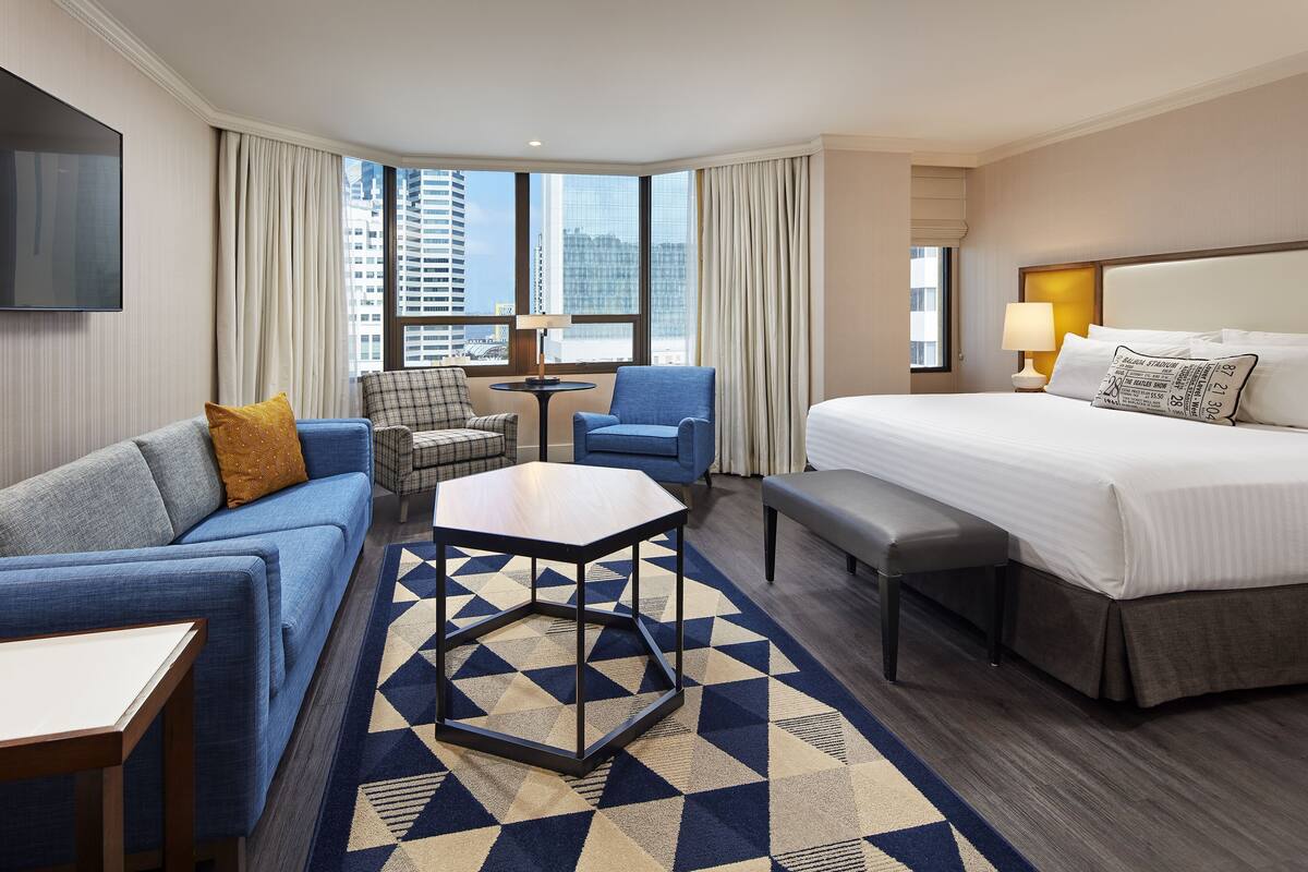A hotel room with a bed, a sofa, a chair, a small table, and a view of the cityscape.