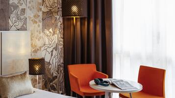A hotel room with a bed, two orange chairs, a small table, and two lamps.