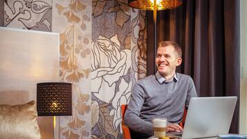 A man sitting at a table with a laptop, a glass of beer, and a plate of food. The room has patterned wallpaper and a decorative lamp.