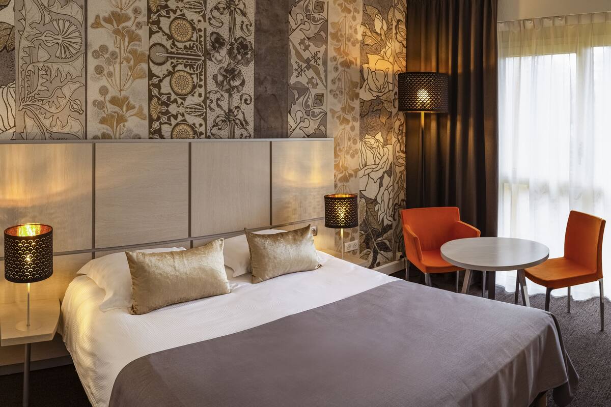 A hotel room with a large bed, two bedside lamps, a small table, two orange chairs, and a patterned wall.