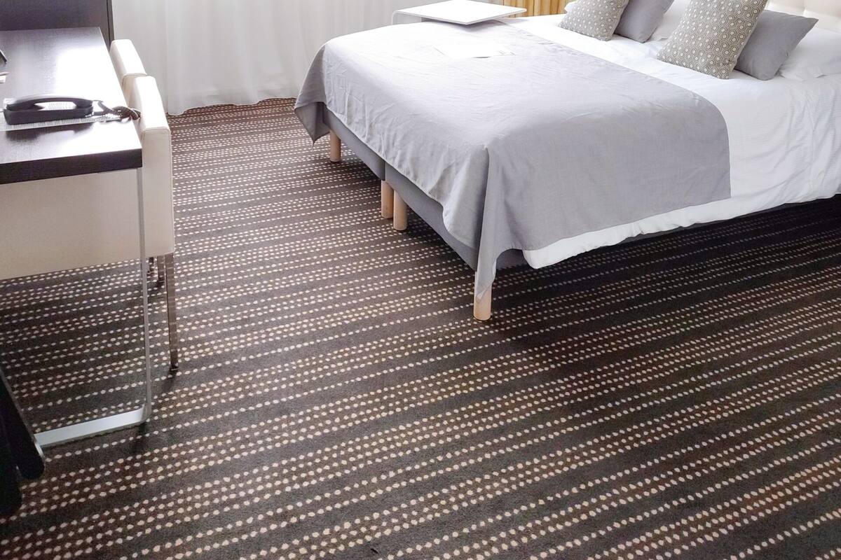 A hotel room with a bed, bedside table, lamp, and a view of the outdoors.
