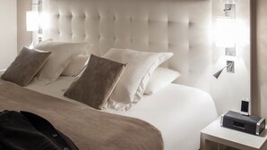 A hotel room with a tufted headboard, a bed with white linens and multiple pillows, a bedside table with a phone and a small electronic device, and a wall-mounted light fixture. - Mercure Bourg En Bresse (Bourg-en-Bresse)
