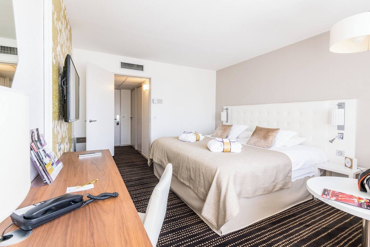 A hotel room with a bed, bedside table, and a desk with a telephone and a magazine.