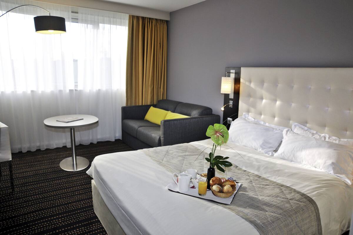 A hotel room with a large bed, a small round table, a grey armchair, and a window with curtains.