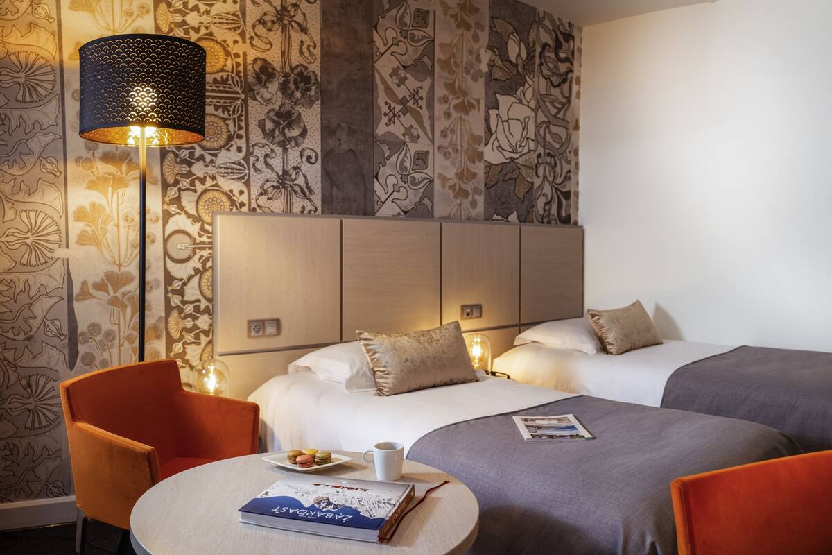 A hotel room with two beds, a round table with a book and a cup, and a lamp.