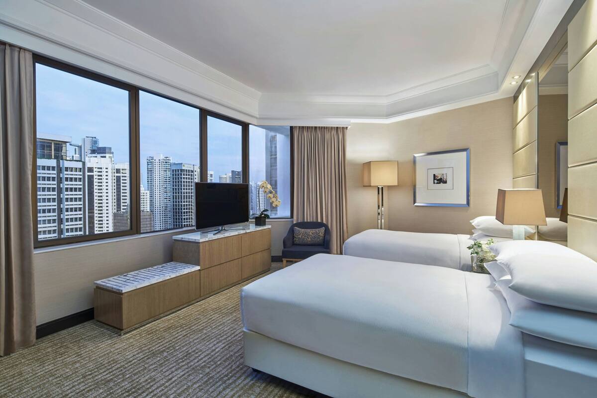 A hotel room with two beds, a TV, a desk, and a large window with a city view.