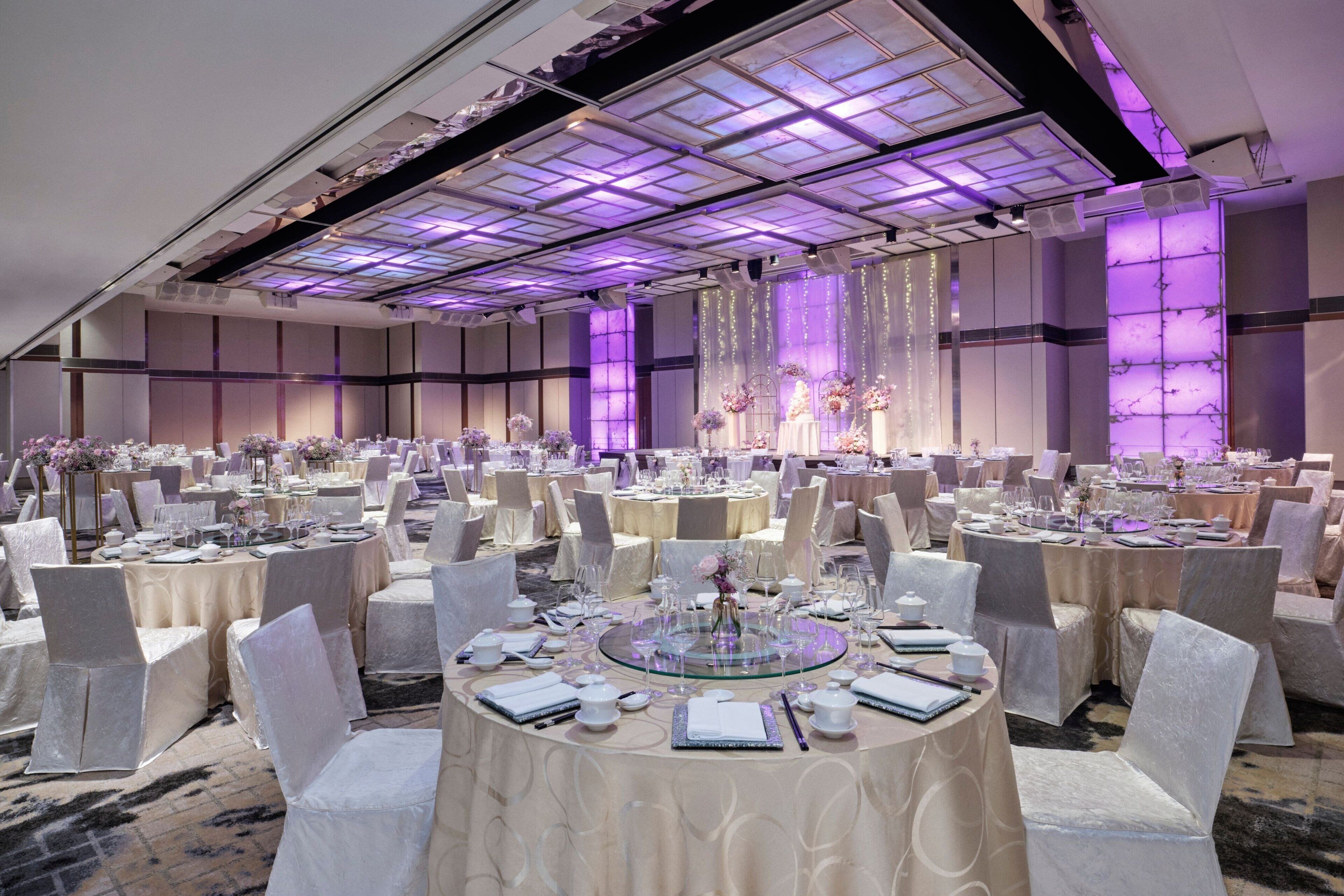 Une vaste salle de banquet, avec des tables rondes disposées pour les événements, dotée d’un plafond haut orné d’un motif en réseau et d’un éclairage d’ambiance.