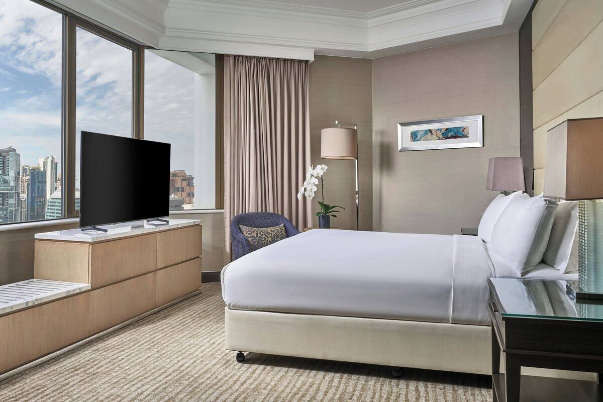 A hotel room with a bed, a TV on a wooden console, a chair, a lamp, and a view of the city through large windows.