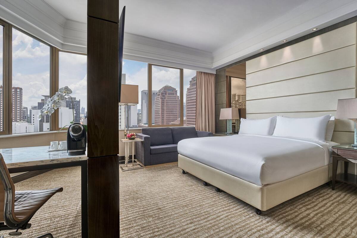 A hotel room with a large bed, a desk with a chair, a sofa, and a view of the cityscape through large windows.
