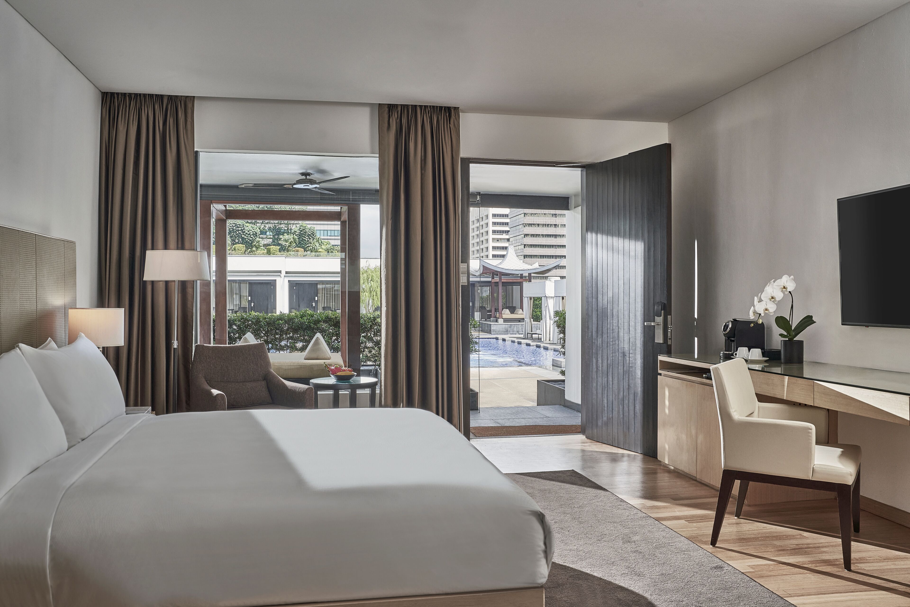 A modern hotel room with a large bed, a desk, a chair, and a view of a swimming pool and buildings.
