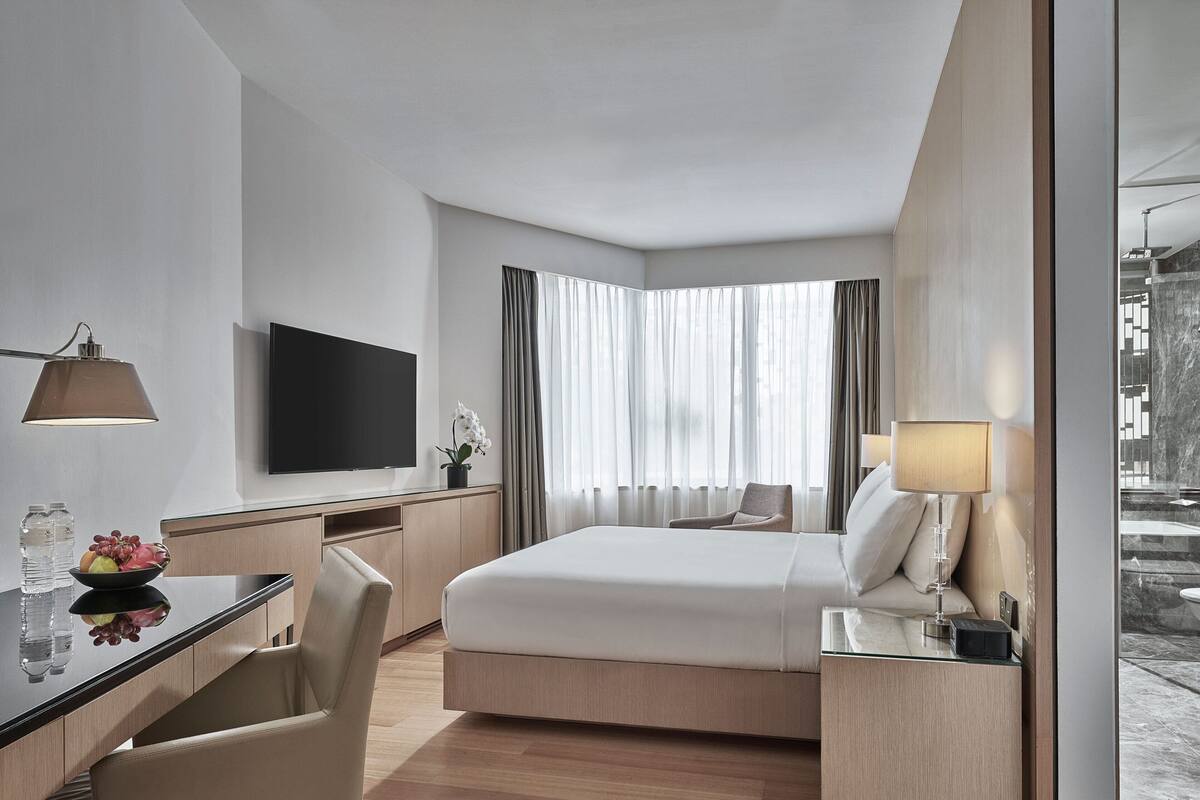 A modern hotel room with a bed, desk, chair, TV, and a view of the outside.