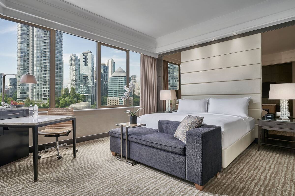 A modern hotel room with a large bed, a desk, and a sofa. The room has a city view through large windows.