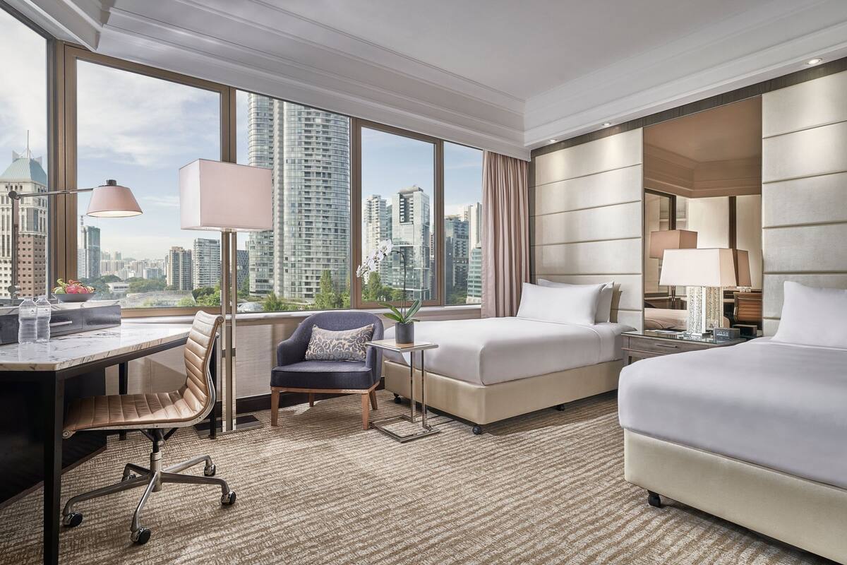 A hotel room with a large window, a desk, a chair, a bed, and a nightstand.