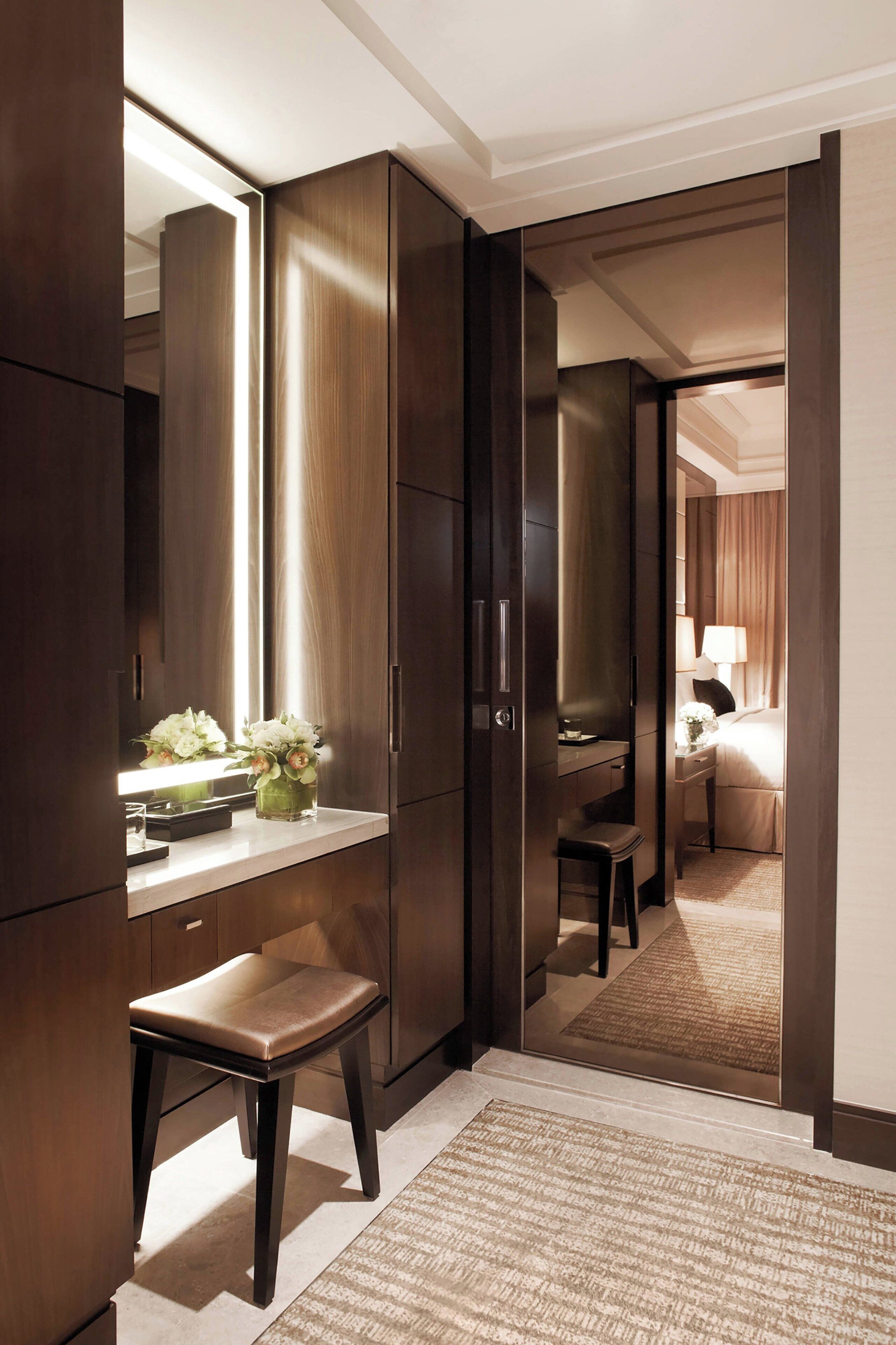 A hotel room with a large wardrobe, a vanity with a mirror, a small stool, and a bed with a nightstand.