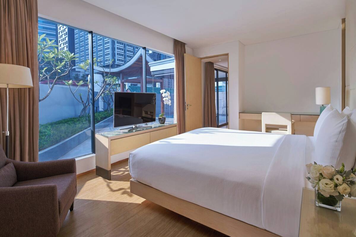 A modern hotel room with a large bed, a desk with a TV, and a view of the cityscape through large windows.