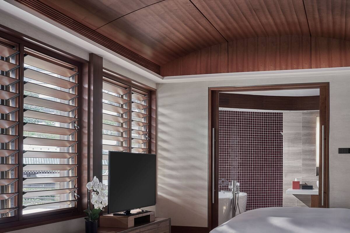 A modern hotel room with a large bed, a TV on a wooden stand, and a view through floor-to-ceiling windows with blinds.