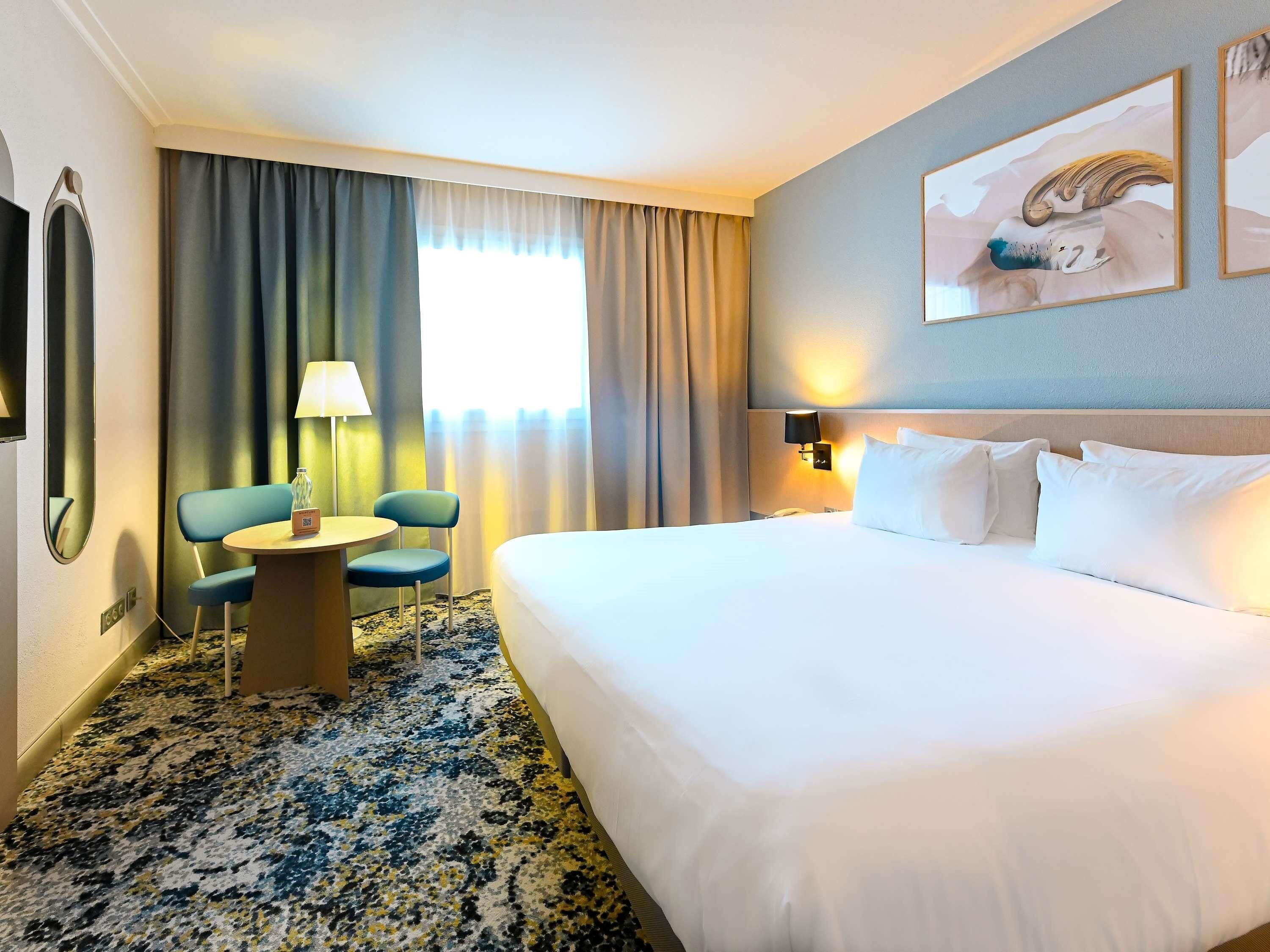 Una habitación de hotel con una cama, dos sillas, una mesita, una lámpara, un televisor montado en la pared y un cuadro.