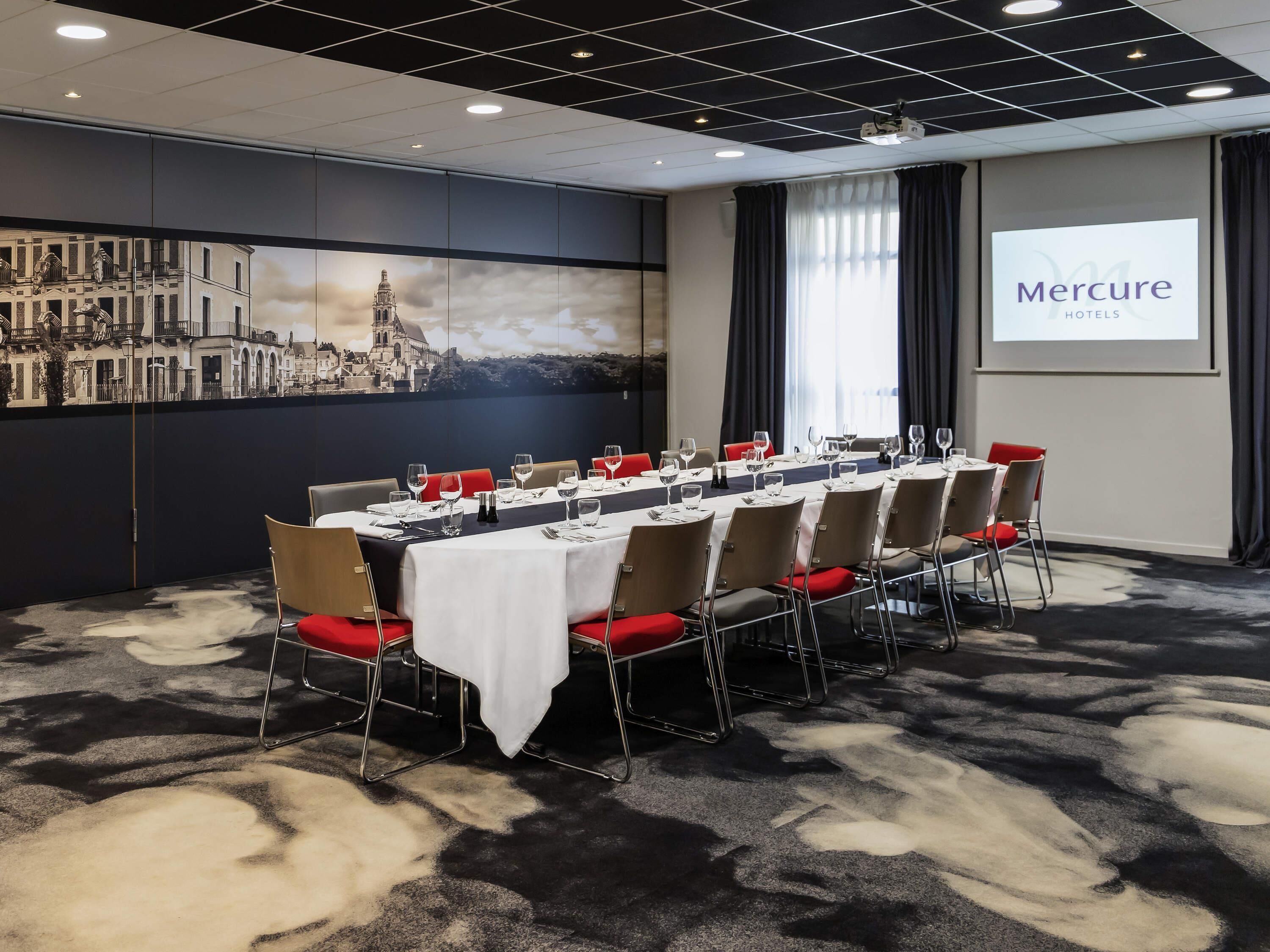 Uma sala de conferências com uma longa mesa preparada para uma reunião, uma tela de projeção exibindo 'Mercure Hotels' e um mural com vista para a cidade.