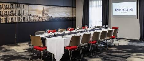 Uma sala de conferências com uma longa mesa preparada para uma reunião, uma tela de projeção exibindo 'Mercure Hotels' e um mural com vista para a cidade.