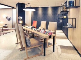 Une pièce moderne et bien éclairée, avec une longue table où sont exposés des magazines, des sièges confortables et un mur foncé à motifs.