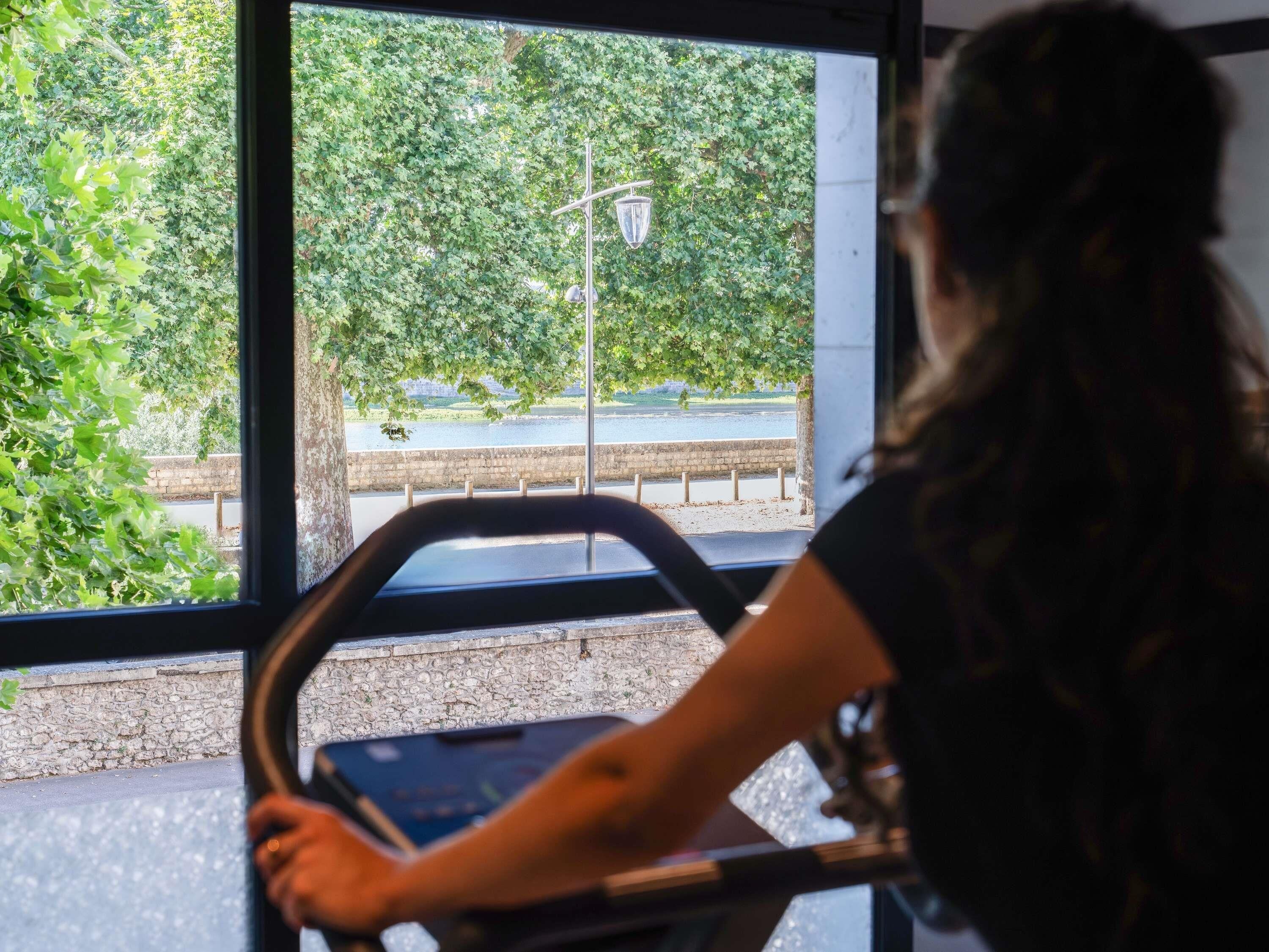 A person using a stationary bike inside a room with large windows overlooking a paved area with trees and a street lamp.