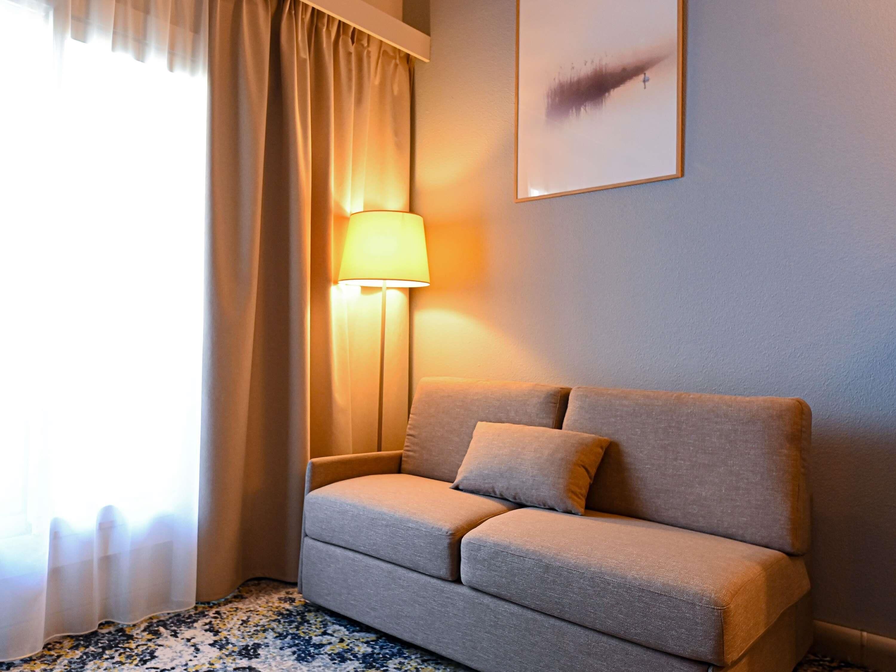 Quarto com sofá, luminária, janela com cortinas e uma obra de arte na parede.