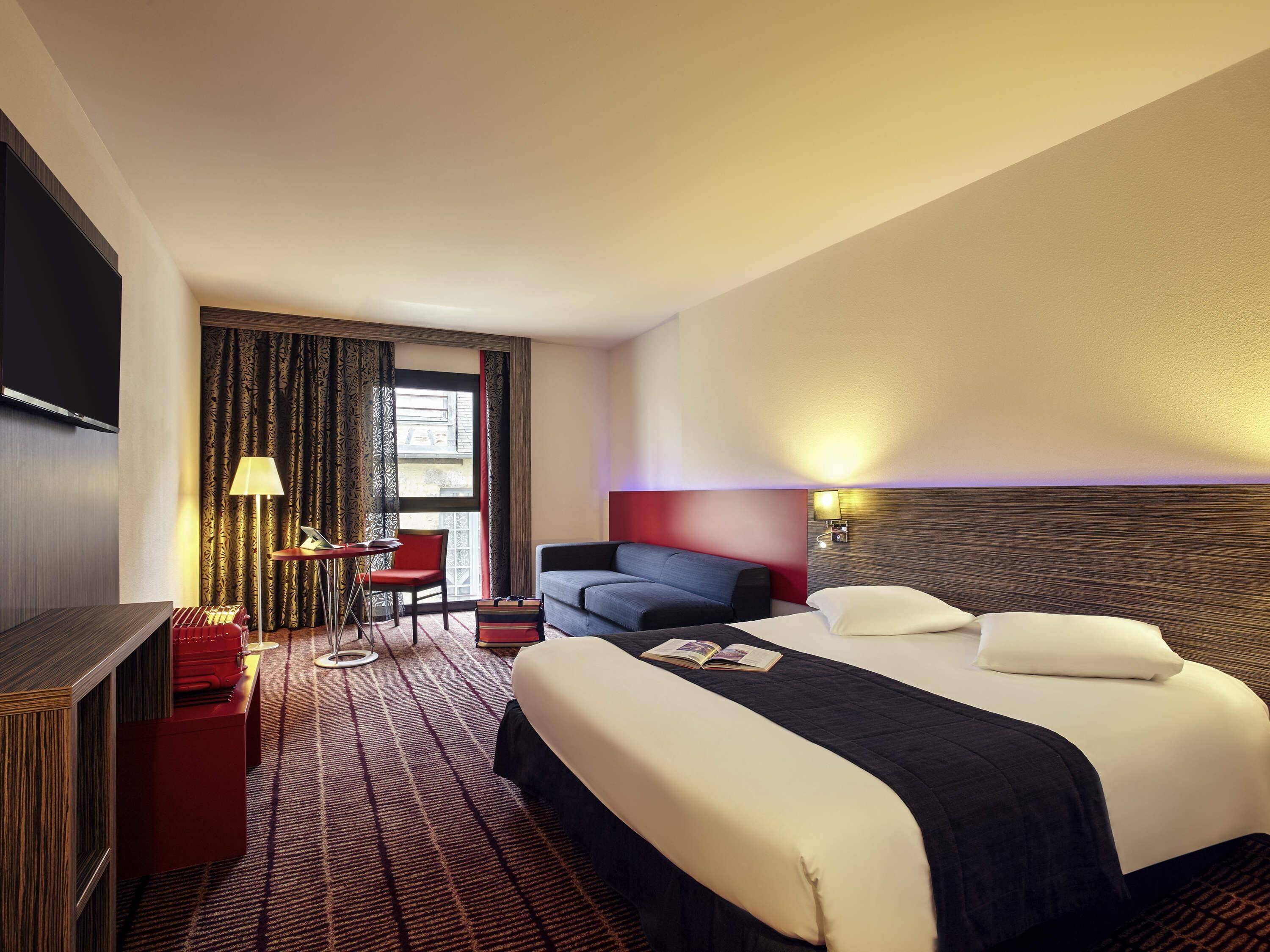A hotel room with a bed, a desk with a chair, a TV, and a window with curtains.
