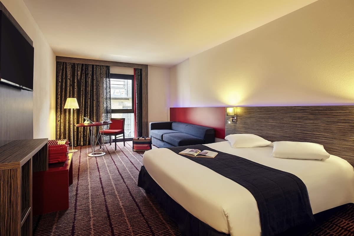 A hotel room with a bed, a desk with a chair, a TV, and a window with curtains.
