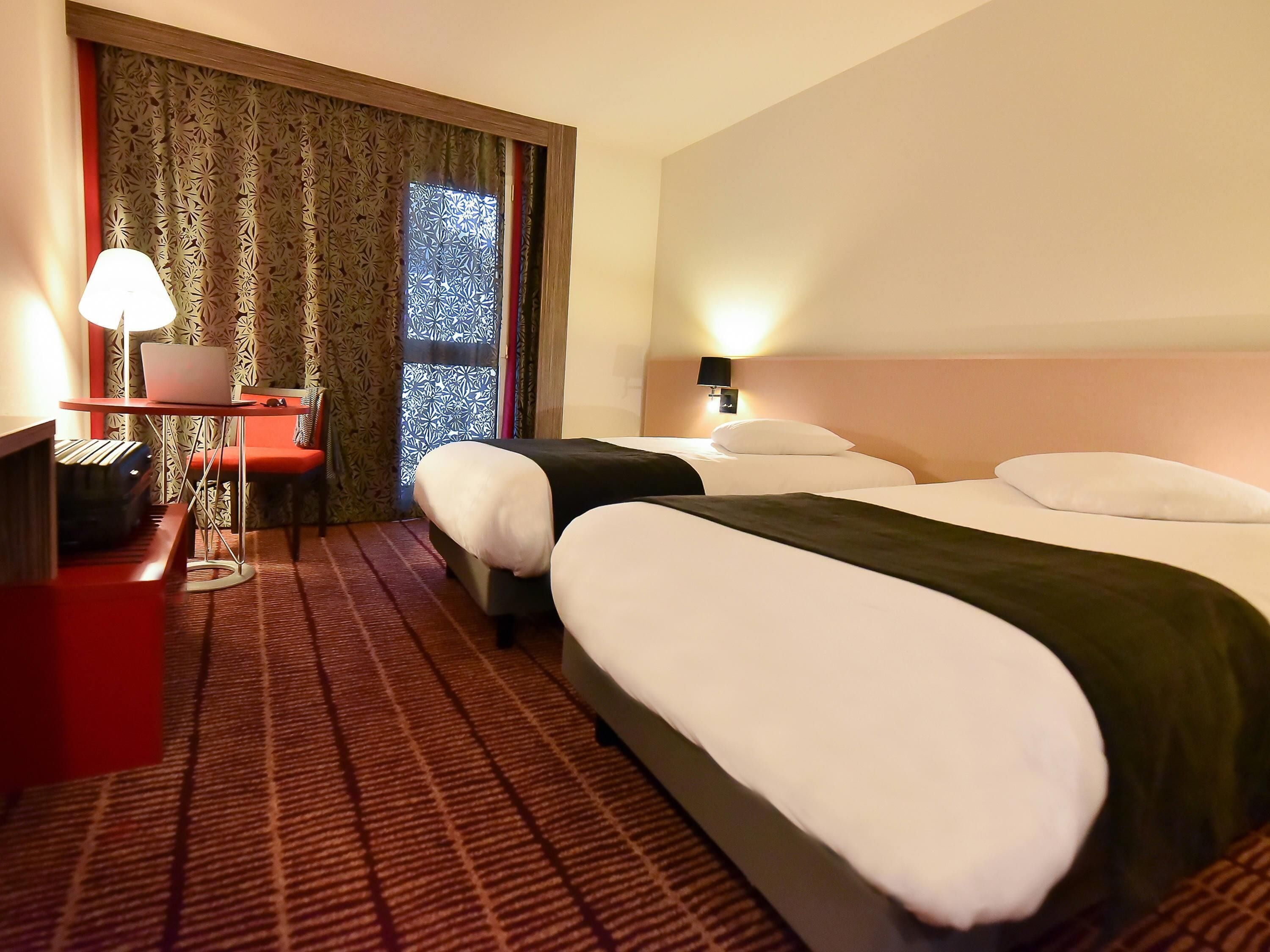 A hotel room with two beds, a desk with a laptop, a lamp, and a window with patterned curtains.