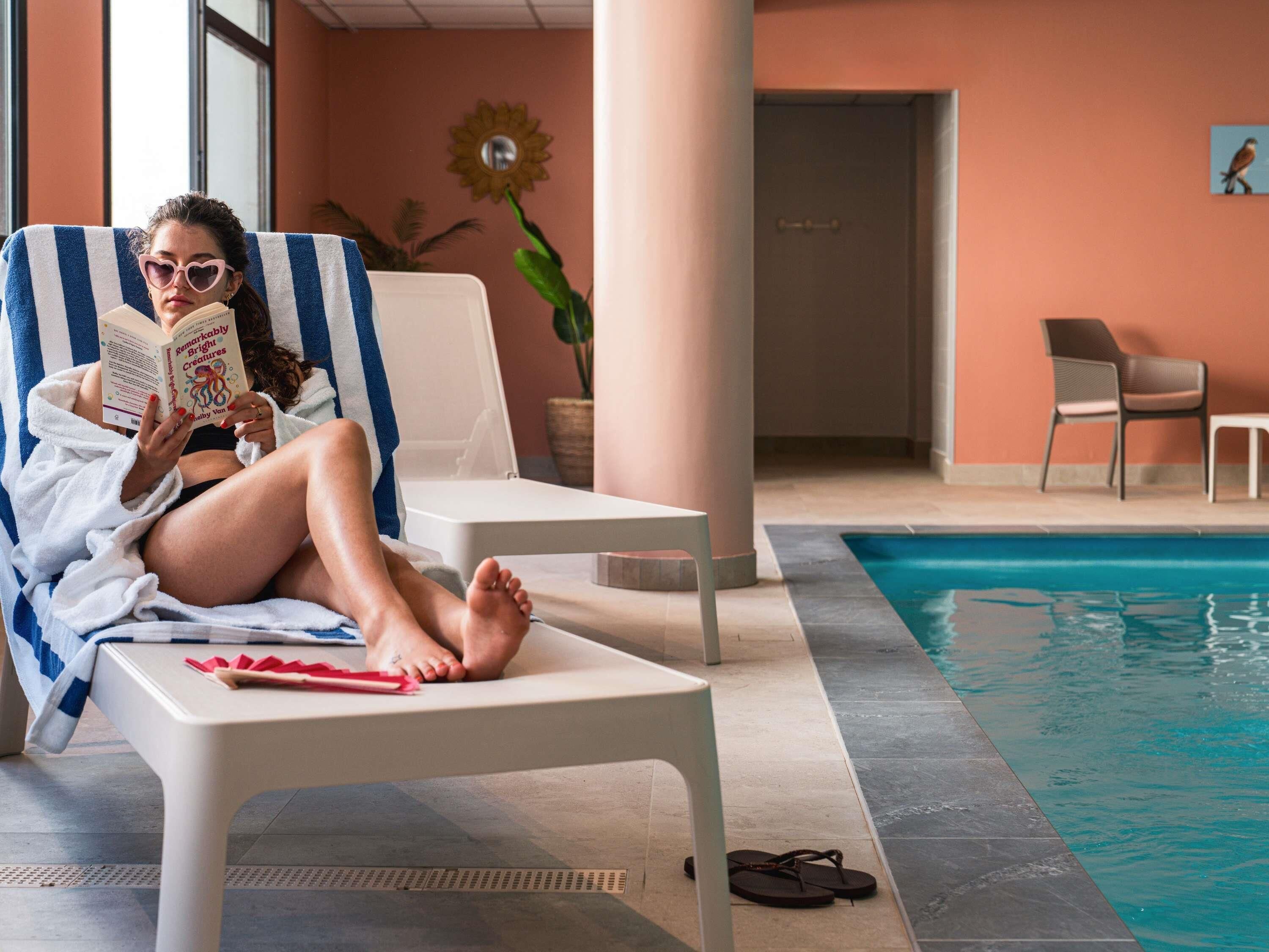 Une personne se détend sur une chaise à rayures, au bord d’une piscine, et lit un livre.