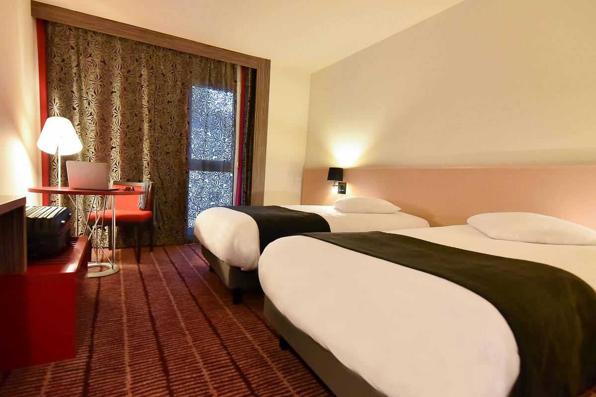 Habitación de hotel con dos camas, un escritorio, una laptop, una lámpara y una ventana con cortinas estampadas.