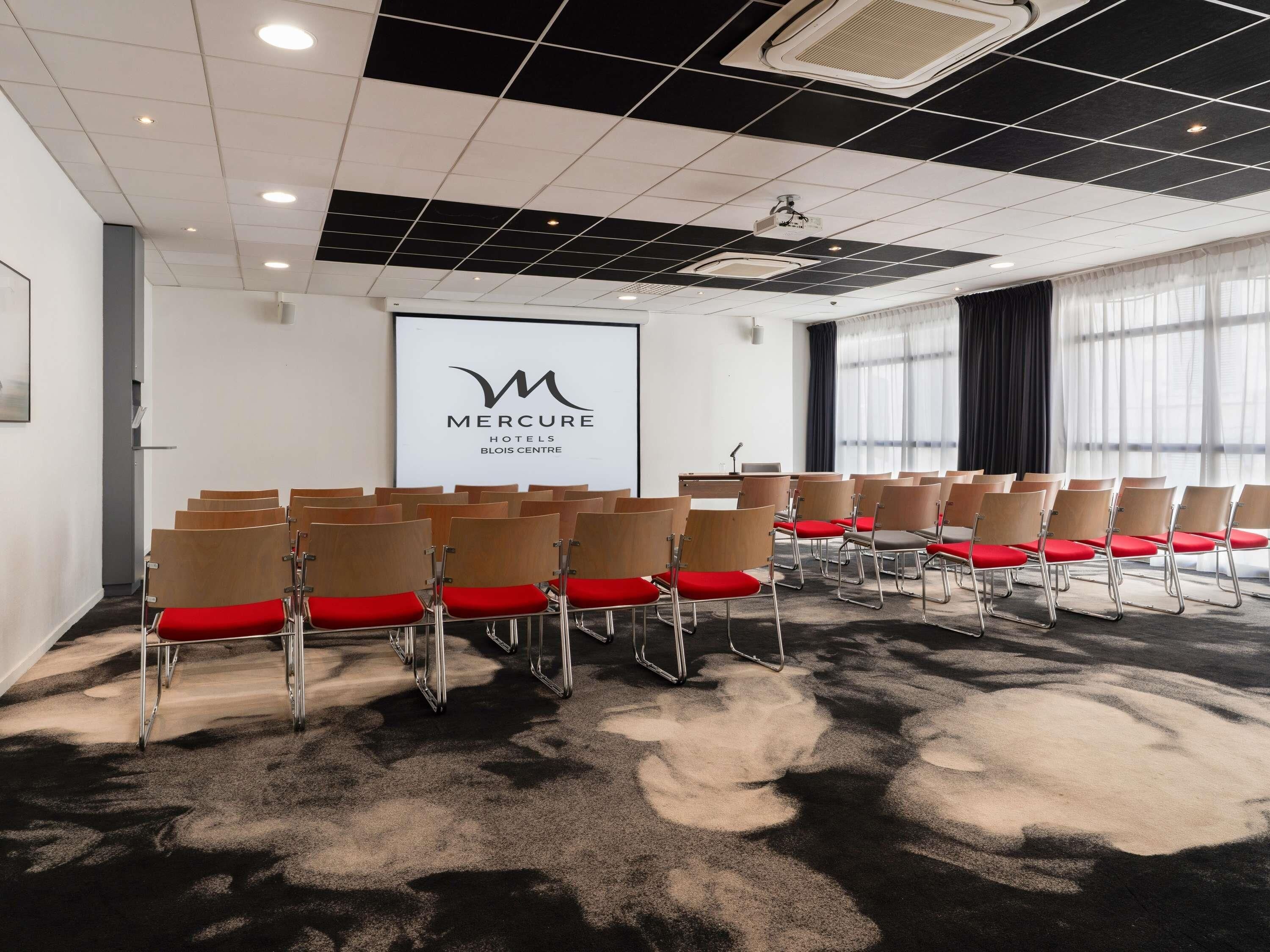 A conference room with rows of chairs, a projector screen, and a logo for Mercure Hotels Blois Centre.