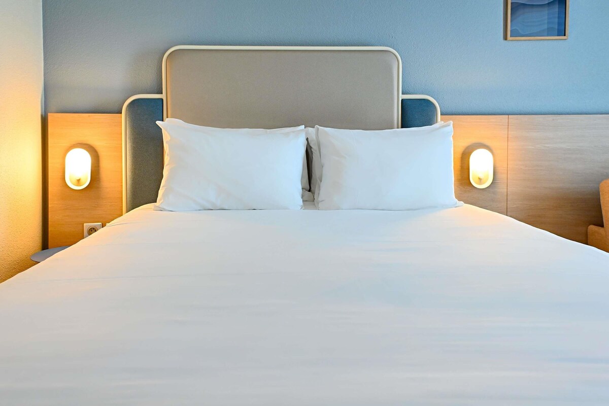 Una cama bien hecha con dos almohadas, cabecera y dos lámparas fijadas a la pared.