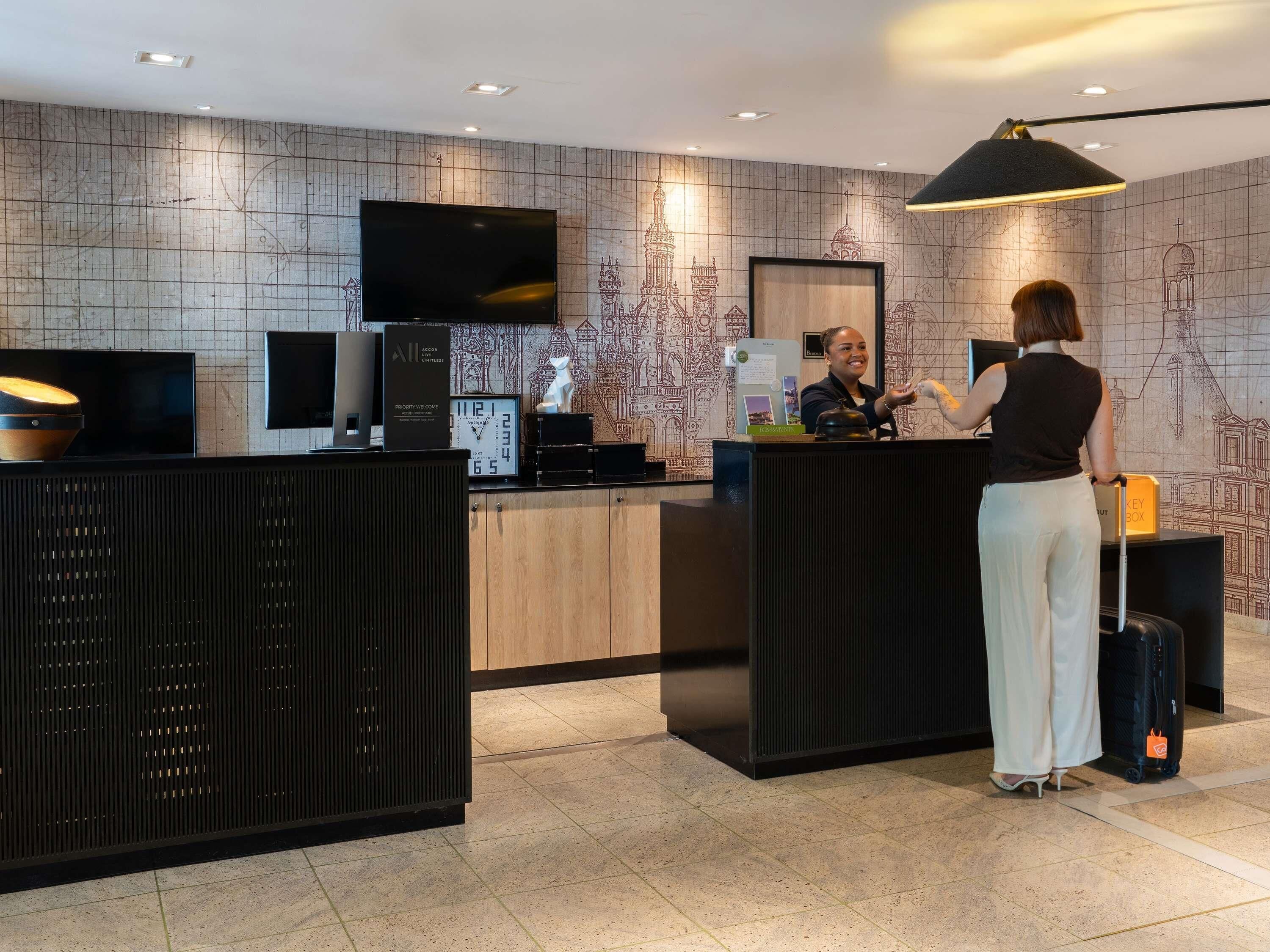 A hotel reception area with a reception desk, a clock, a television, and a wall with architectural drawings.