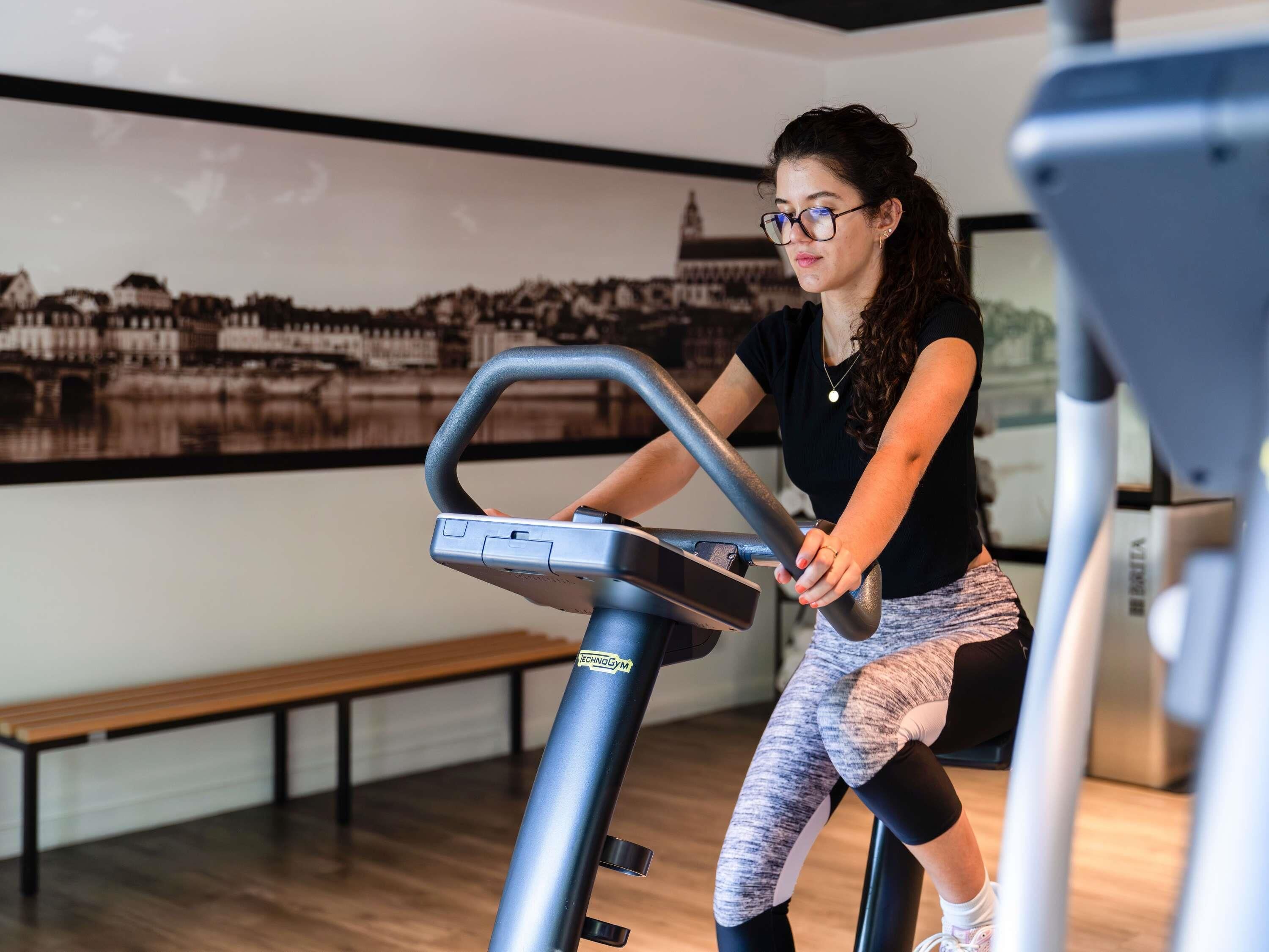 Une personne fait de l’exercice sur un vélo d’appartement dans une pièce où se trouvent un banc et un tableau accroché au mur.